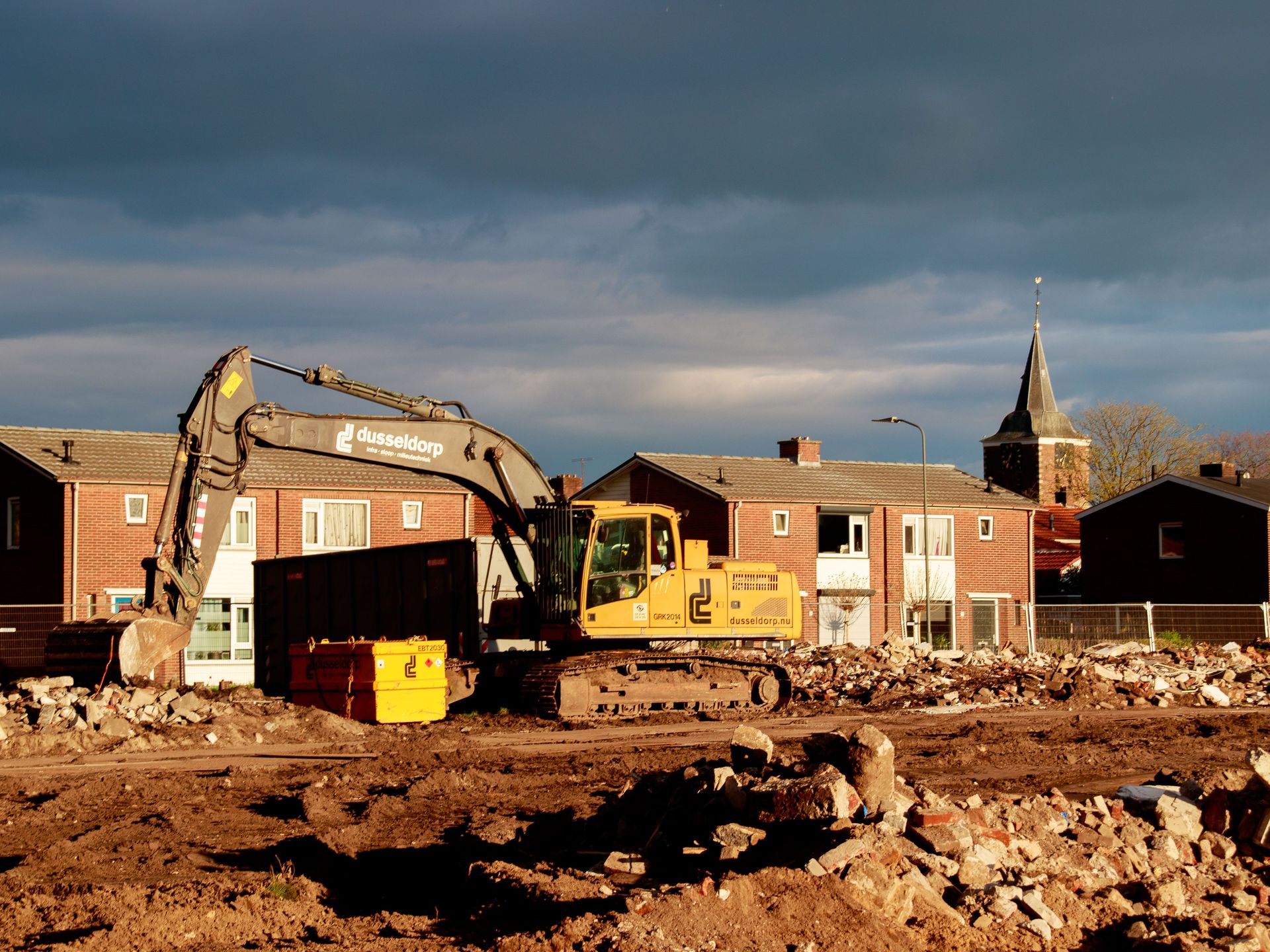 Excavator at a construction site near residential houses under a dramatic sky.