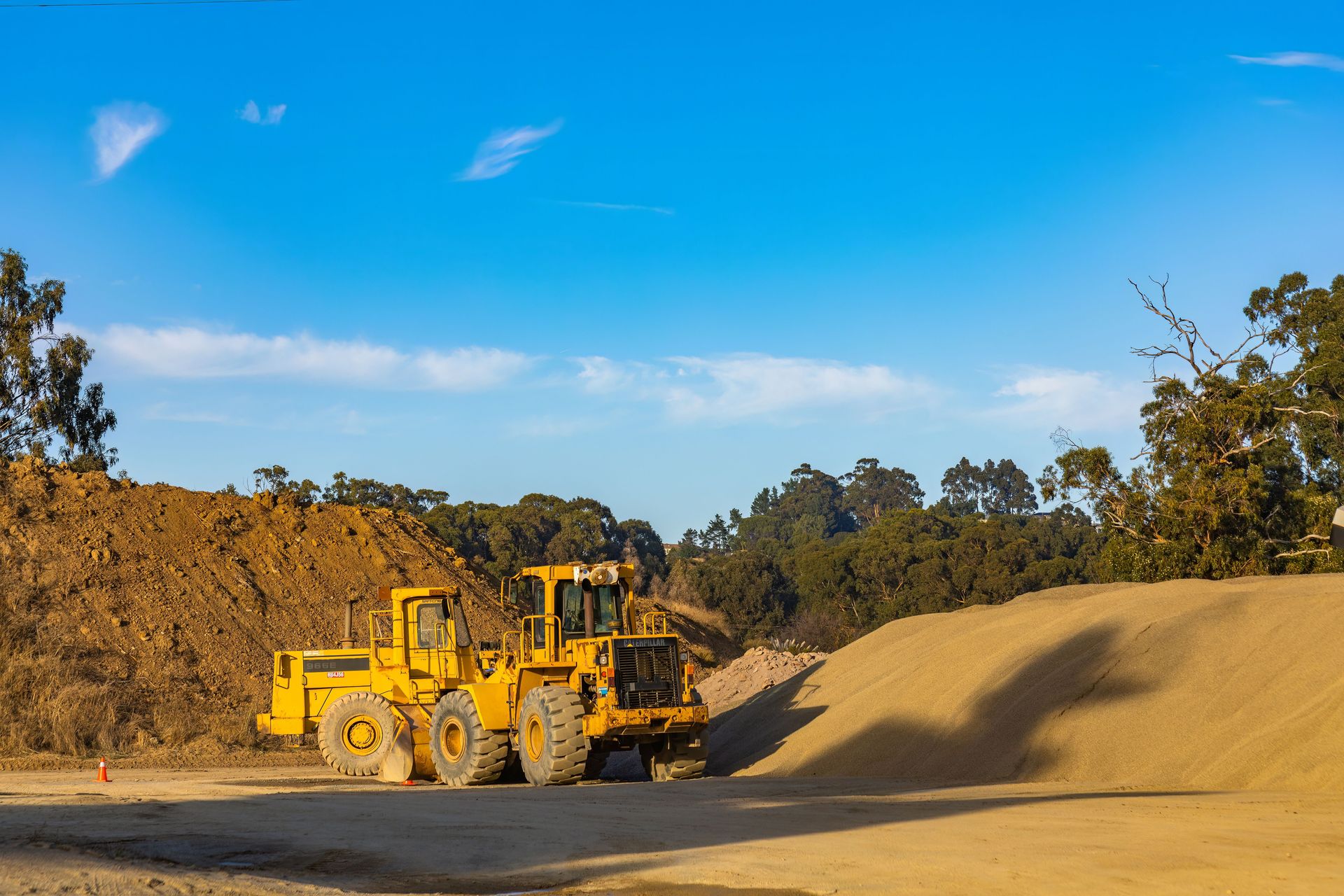 A large yellow tractor at a construction site with gravel and sand piles under a clear blue sky.