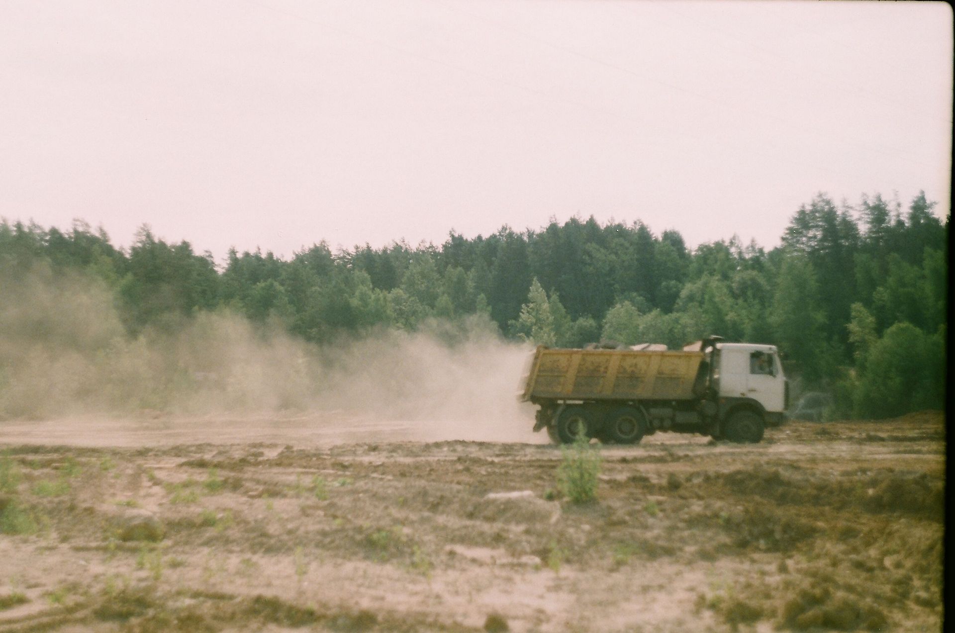 A dump truck kicks up dust on a rural site surrounded by trees.